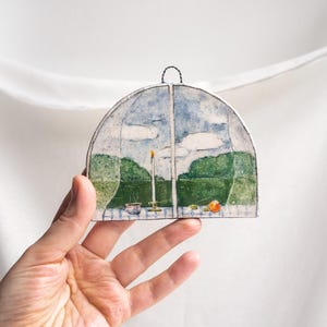 May include: A semi-circular ceramic wall hanging featuring a painted landscape scene of a green field, blue sky with white clouds, a lit candle, a small bowl, and an orange fruit on a table, finished with a dark wire loop for hanging.