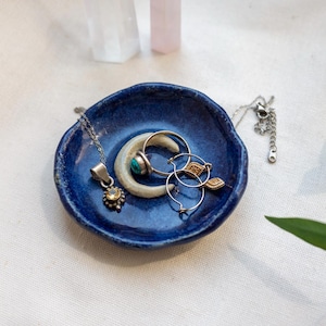 Puede incluir: Un pequeño plato de cerámica hecho a mano, de forma redonda, con un esmalte brillante de color azul intenso. Dentro del plato hay un objeto blanco en forma de media luna, un collar de plata con un colgante y un par de pendientes de aro de plata.
