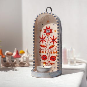 May include: A tall, arched ceramic candle holder with a light blue exterior and a cream-colored interior. The interior features a hand-painted floral design in red, orange, and black, with a small lit tea light candle resting at the base.
