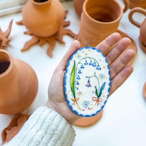Pode incluir: Uma placa de cerâmica oval com superfície branca texturizada, apresentando uma ilustração pintada à mão de flores azuis e verdes em forma de sino, pequenas estrelas coloridas e laços laranja, com uma borda azul recortada e um gancho metálico.