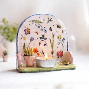 Pode incluir: Um castiçal de cerâmica com um design floral. O fundo arqueado é decorado com flores e vegetação pintadas. Uma pequena suculenta em vaso e uma vela de chá acesa estão na base. O suporte é pintado com uma borda azul.