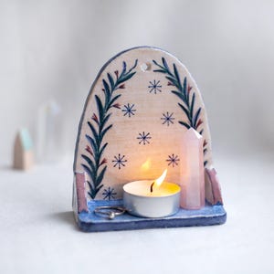 Könnte beinhalten: Ein Kerzenhalter aus Keramik mit blauem Sockel und gewölbtem Oberteil, verziert mit grünen und roten Blumenmustern und blauen Sternakzenten. Eine brennende Teelichtkerze und ein rosa Kristall befinden sich im Inneren. Ein silberner Ring liegt auf dem Sockel.