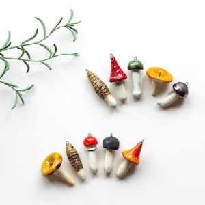 Pode incluir: Uma coleção de ornamentos de cogumelos feitos à mão em várias cores, incluindo vermelho, amarelo e castanho. Os ornamentos têm caules brancos e estão dispostos sobre uma superfície branca. Um ramo verde e frondoso está no canto superior esquerdo.