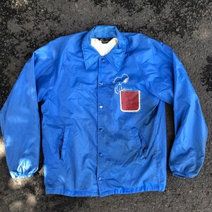 Puede incluir: Chaqueta azul abotonada con un gráfico de Snoopy en el bolsillo delantero. La chaqueta tiene cuello, mangas largas y forro blanco. El gráfico de Snoopy es en blanco y negro, con un bolsillo rojo.