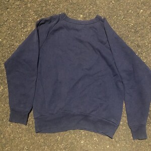 May include: A navy blue sweatshirt with a crew neck and long sleeves. The sweatshirt is made of a soft material and has ribbed cuffs and hem. The garment is laid flat on a textured surface.
