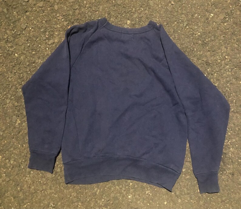 May include: A navy blue sweatshirt with a crew neck and long sleeves. The sweatshirt is made of a soft material and has ribbed cuffs and hem. The garment is laid flat on a textured surface.