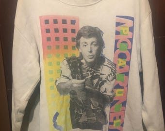 Puede incluir: Sudadera vintage de color crema con una imagen en blanco y negro de un músico sosteniendo una guitarra. El gráfico incluye formas geométricas coloridas y el nombre "MCCARTNEY" en texto vertical. La sudadera tiene cuello redondo y mangas largas.