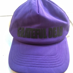 May include: Purple trucker hat with a black mesh back and the words "Grateful Dead" in black lettering on the front.