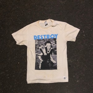 Può includere: Una maglietta bianca con una foto in bianco e nero di una persona con un ciuffo e la parola "DESTROY" stampata in blu sopra la foto.