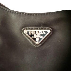 May include: Close-up of a black nylon handbag with a silver triangular logo that reads "PRADA" in all caps.