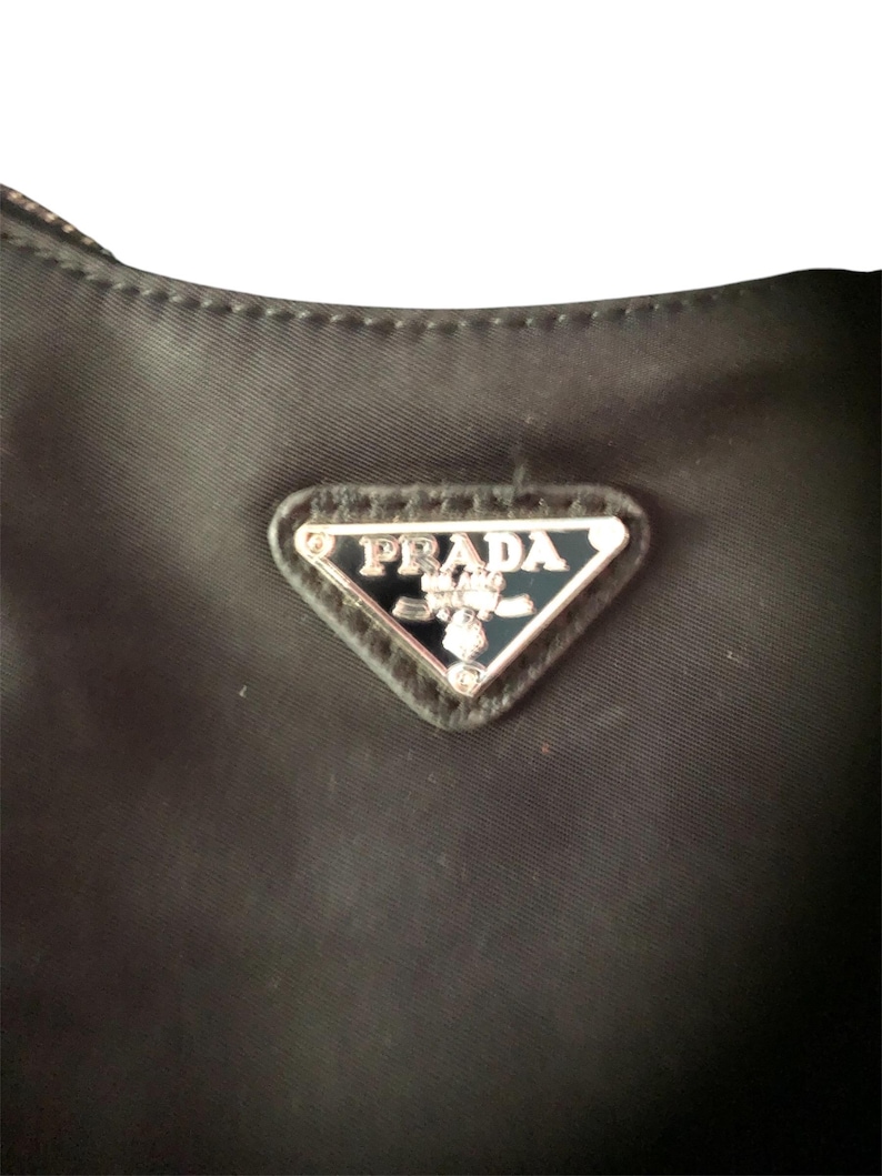 May include: Close-up of a black nylon handbag with a silver triangular logo that reads "PRADA" in all caps.