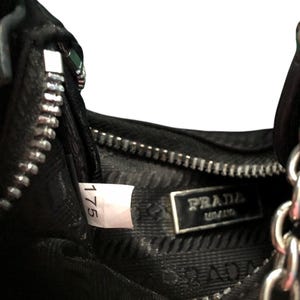May include: Close-up of the inside of a black leather handbag with a silver zipper and a Prada label. The label has the text "PRADA" and "MILANO" on it. A white tag with the number "175" is visible.