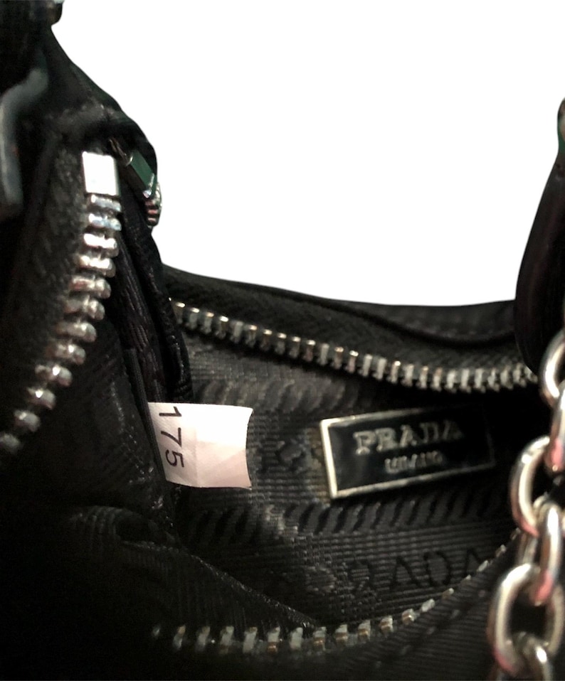 May include: Close-up of the inside of a black leather handbag with a silver zipper and a Prada label. The label has the text "PRADA" and "MILANO" on it. A white tag with the number "175" is visible.