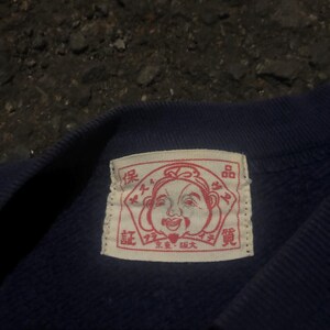May include: Close-up of a navy blue garment with a rectangular label. The label features a red illustration of a smiling face and Japanese text. The garment appears to be made of a soft, textured fabric.
