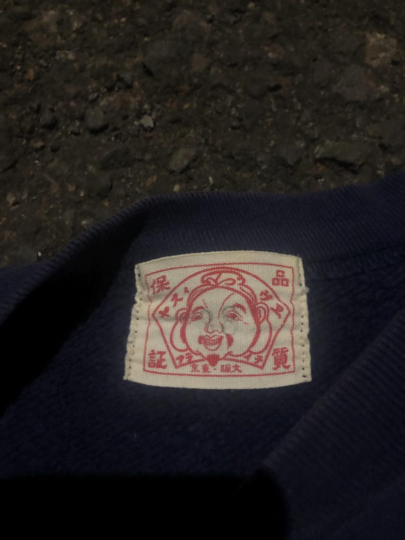 May include: Close-up of a navy blue garment with a rectangular label. The label features a red illustration of a smiling face and Japanese text. The garment appears to be made of a soft, textured fabric.