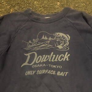 May include: Navy blue sweatshirt with a gray graphic of a fisherman in a boat catching a fish. The text "Dowluck OSAKA-TOKYO ONLY SURFACE BAIT" is printed below the graphic. The sweatshirt has a crew neck and raglan sleeves.
