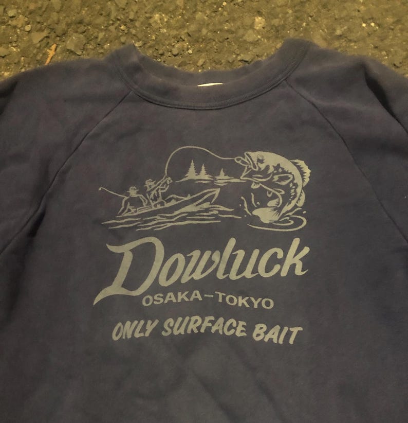 May include: Navy blue sweatshirt with a gray graphic of a fisherman in a boat catching a fish. The text "Dowluck OSAKA-TOKYO ONLY SURFACE BAIT" is printed below the graphic. The sweatshirt has a crew neck and raglan sleeves.