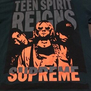 以下が含まれることがあります： バンドの黒とオレンジのグラフィックプリントが施されたターコイズブルーのTシャツ。グラフィックには「Teen Spirit Reigns」と「Supreme」の文字が書かれています。