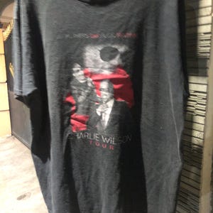 May include: Black t-shirt featuring a graphic of Charlie Wilson in a suit, with the text "CHARLIE WILSON TOUR" in red. The shirt also has the text "IN LOVERS'S MUSIC" in white. The shirt is made of a soft material.