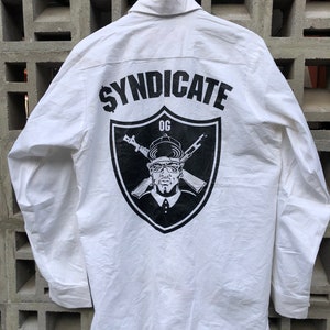 May include: White long-sleeve button-down shirt with a black and white graphic on the back. The graphic features a shield with the text "Syndicate OG" and a cartoon character wearing a hat and sunglasses.