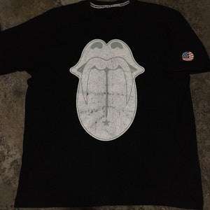 Puede incluir: Camiseta negra con un gráfico blanco de una lengua con dientes. La camiseta tiene un pequeño parche de la bandera estadounidense en la manga derecha.