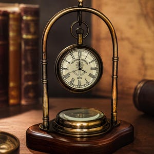 May include: A brass desk clock with a circular face featuring Roman numerals, the text "VICTORIA LONDON" and "1876", and a central anchor graphic. The clock hangs from a brass arch mounted on a dark wooden base, which also holds a circular compass.