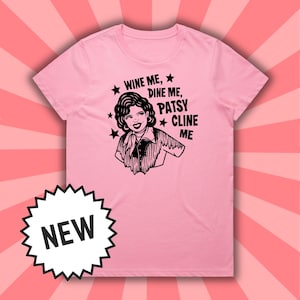 Puede incluir: Una camiseta rosa claro con una ilustración en blanco y negro de una mujer y el texto "WINE ME, DINE ME, PATSY CLINE ME" con acentos de estrellas. Una pegatina "NUEVO" está en la esquina inferior izquierda.