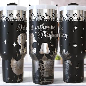 May include: Black stainless steel tumbler with a white lid and handle. The tumbler features a silver design with the text "I'd rather be thrifting" and illustrations of a goose, a shopping cart, and plants. The design includes stars and floral accents.