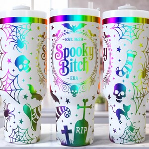 May include: White tumbler with a Halloween theme, featuring a rainbow gradient rim and handle. The design includes spiders, cobwebs, bats, skulls, and the text "Spooky Bitch - ERA" in a colorful, circular frame. The tumbler has a white lid.