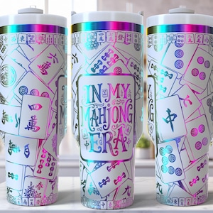 May include: White tumbler with a wraparound design featuring colorful Mahjong tiles and the text "IN MY MAHJONG ERA." The tumbler has a white handle and lid with a rainbow iridescent trim. The design includes Chinese characters and colorful accents.