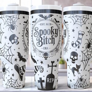 May include: White tumbler with a black Halloween-themed design. The design includes spiderwebs, spiders, bats, skulls, and the text "Spooky Bitch". The tumbler has a white handle and lid, perfect for spooky season.