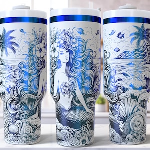 May include: White travel mug with a wraparound design featuring a mermaid, ocean waves, palm trees, and fish in shades of blue. The mug has a white handle and a blue accent band at the top.