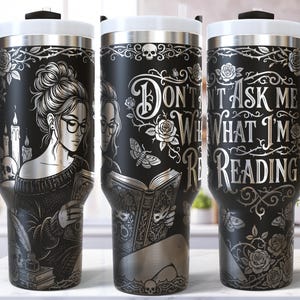 May include: Black travel mug with a white lid and handle, featuring an engraved design. The design includes a woman reading, skulls, roses, and the text "Don't Ask Me What I'm Reading". The mug is approximately 23 cm tall.