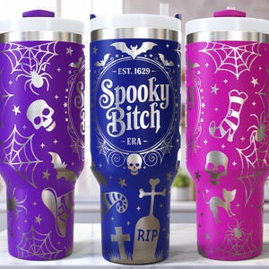 May include: Three Halloween-themed travel mugs in purple, blue, and pink. Each mug features a white lid, handle, and decorative silver spiderwebs, bats, and skulls. The blue mug has the text "Spooky Bitch".