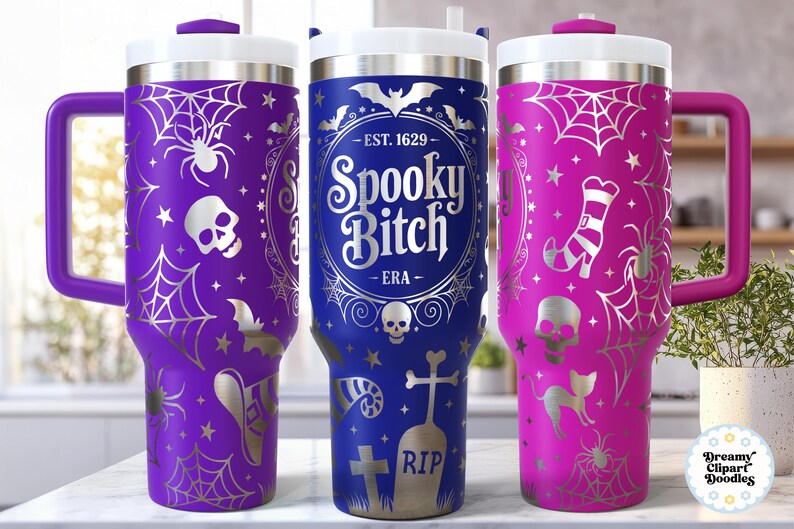May include: Three Halloween-themed travel mugs in purple, blue, and pink. Each mug features a white lid, handle, and decorative silver spiderwebs, bats, and skulls. The blue mug has the text "Spooky Bitch".