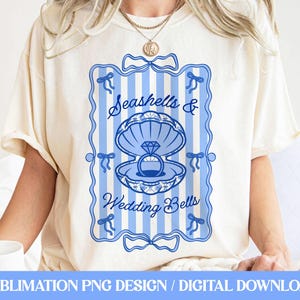 Könnte beinhalten: Cremefarbenes T-Shirt mit blau-weißem Streifendesign. Das Design zeigt die Worte "Seashells & Wedding Bells" und eine Muschel mit einem Diamantring. Auf der Unterseite des Shirts steht "SUBMLIMATION PNG DESIGN / DIGITAL DOWNLOAD."