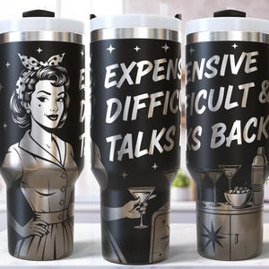 May include: Black travel mug with a white lid and handle. The mug features a retro illustration of a woman and the text "Expensive Difficult & Talks Is Back". The design includes cocktail glasses.