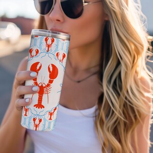 May include: A white tumbler with a red lobster design and blue accents. The stainless steel travel mug has a silver lid and is cylindrical in shape. The lobster pattern is repeated around the tumbler's surface.