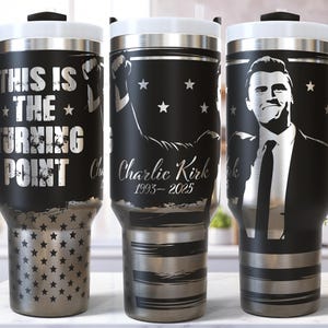 May include: Black and silver travel mugs with handles. One mug has the text "THIS IS THE TURNING POINT". Another mug has a graphic of a person with the text "Charlie Kirk 1993-2025". The third mug has a portrait of a man.