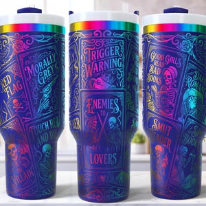 May include: A set of three insulated tumblers with a dark blue background and a wraparound design featuring book-themed graphics. The text includes "Trigger Warning", "Enemies to Lovers", and "Good Girls Read Bad Books". Each tumbler has a blue handle and a rainbow-colored rim.