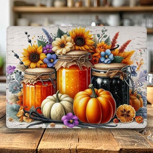 Può includere: Un tagliere decorativo con tre barattoli di vetro pieni di conserve, circondati da girasoli, zucche e foglie autunnali. I barattoli contengono conserve arancioni e nere. Lo sfondo è in legno bianco invecchiato.