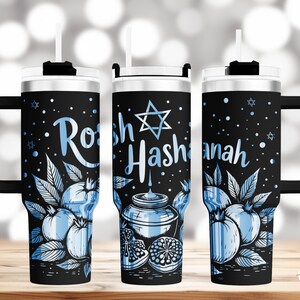 Pode incluir: Uma caneca de viagem de aço inoxidável preto com alça e um canudo azul. A caneca tem um design de romãs, uma Estrela de Davi e o texto "Rosh Hashanah".