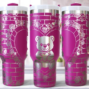 May include: A pink insulated tumbler with a white lid and handle. The tumbler features a silver design of a teddy bear holding a heart, surrounded by a heart-shaped floral arrangement and brick pattern. The tumbler is approximately 10 inches tall.