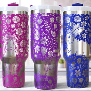 May include: Three stainless steel travel mugs with floral and bunny designs. The mugs are in shades of pink, purple, and blue, with silver accents. Each mug has a handle and a white lid. The bunny is wearing a floral crown.
