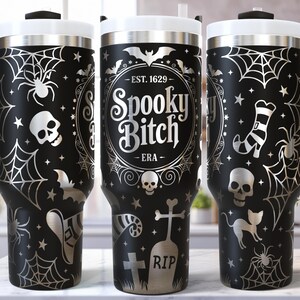 May include: Black travel mug with a white lid and handle, featuring a Halloween-themed design. The design includes spiderwebs, spiders, bats, skulls, and the text "Spooky Bitch - ERA".