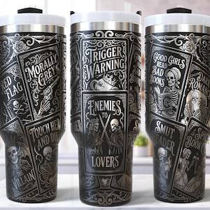 Puede incluir: Taza de viaje negra con tapa y asa blancas, con un diseño gótico. La taza está adornada con ilustraciones de calaveras, naipes y frases como "Trigger Warning" y "Good Girls Read Bad Books."