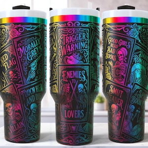 May include: Three black travel mugs with a wraparound design featuring gothic illustrations and text. The text includes "Trigger Warning," "Enemies to Lovers," and "Good Girls Read Bad Books." The mugs have a black handle and a white lid with a rainbow band.