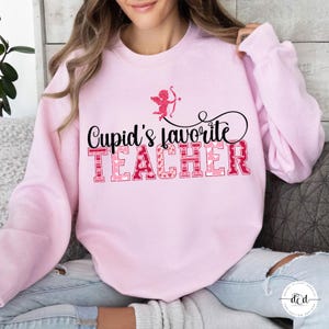May include: A pale pink sweatshirt featuring the phrase "Cupid's Favourite Teacher" in pink and red lettering. A small Cupid graphic with a bow and arrow is above the text. The sweatshirt is a Valentine's Day themed item.