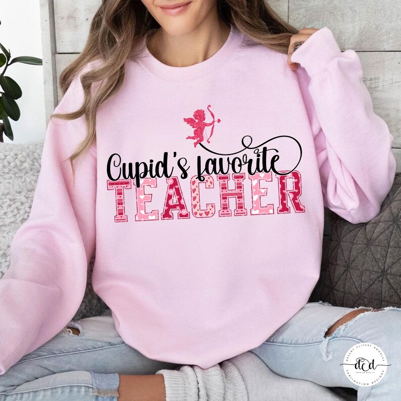 May include: A pale pink sweatshirt featuring the phrase "Cupid's Favourite Teacher" in pink and red lettering. A small Cupid graphic with a bow and arrow is above the text. The sweatshirt is a Valentine's Day themed item.