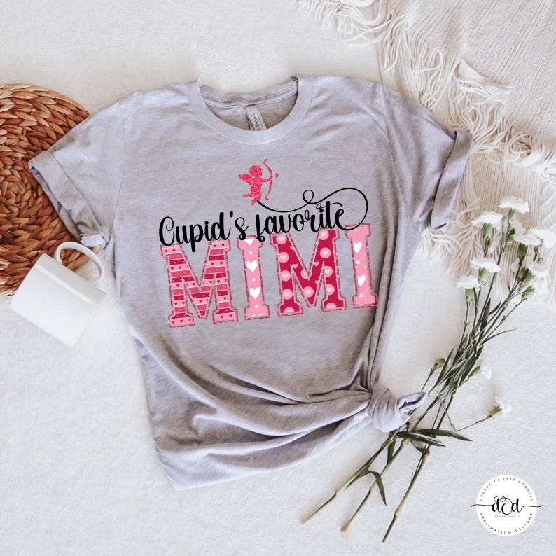 May include: A light grey t-shirt featuring the text "Cupid's favourite MIMI" in black and pink. The word "MIMI" is in a pink, patterned font. A pink Cupid graphic is above the text. The shirt is knotted at the bottom.